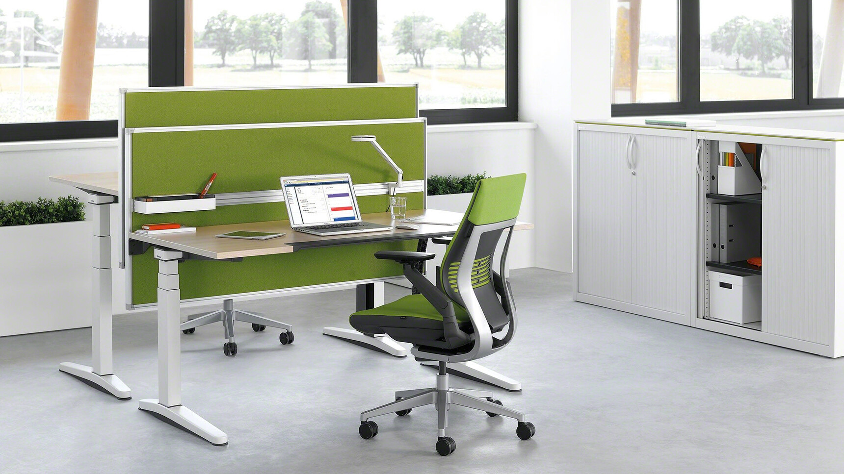 Two Steelcase Gesture Wrapped Back Office Chairs In Green Upholstery And Black Grey Frame And Grey Base Finish On Castors And Black Arms At Two Height Adjustable Desks With Wood Table Top Finish And White Legs