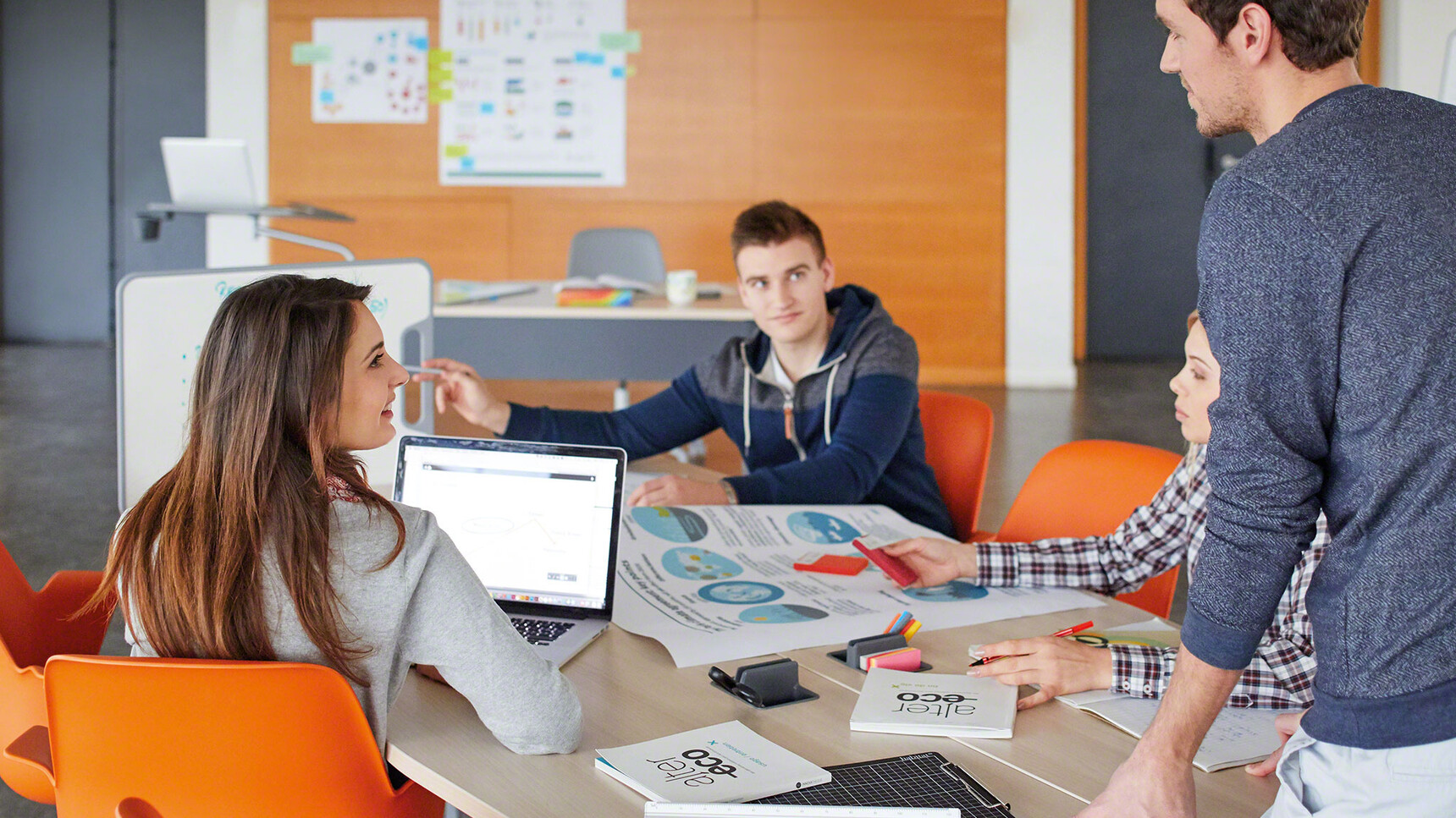 Two Steelcase Verb Rectangular Desks With Wood Table Top Finish And Four Node Chairs With Orange Plastic Seat And Backrest At Desk With Verb Rectangular Desk With Light Wood Finish And Modesty Panel In Use