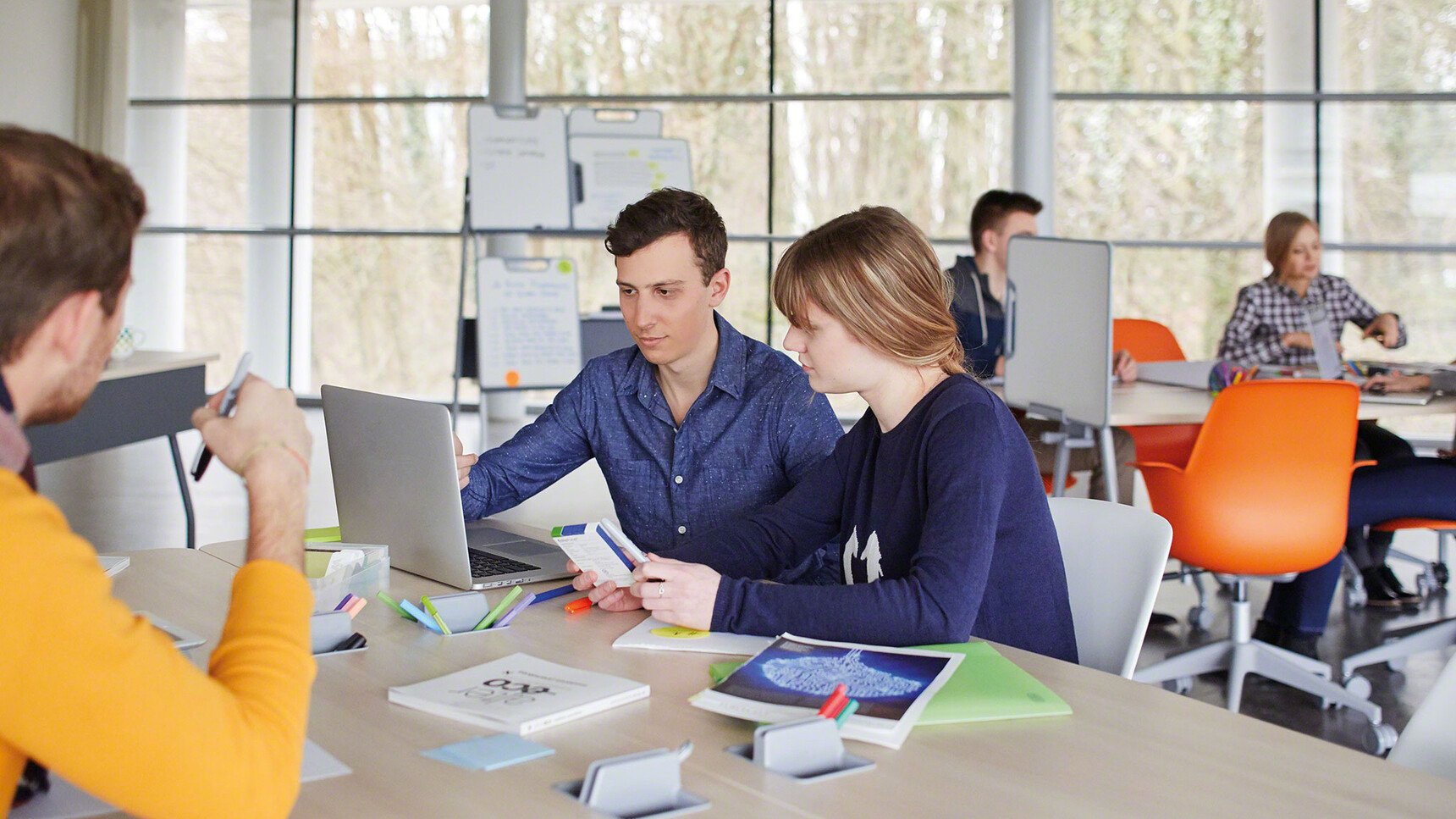Four Steelcase Verb Rectangular Desks With Wood Table Top Finish And Grey Leg Finish With Eight Node Chairs In Orange And White Finish And Grey Base At Desks In Use By Six People