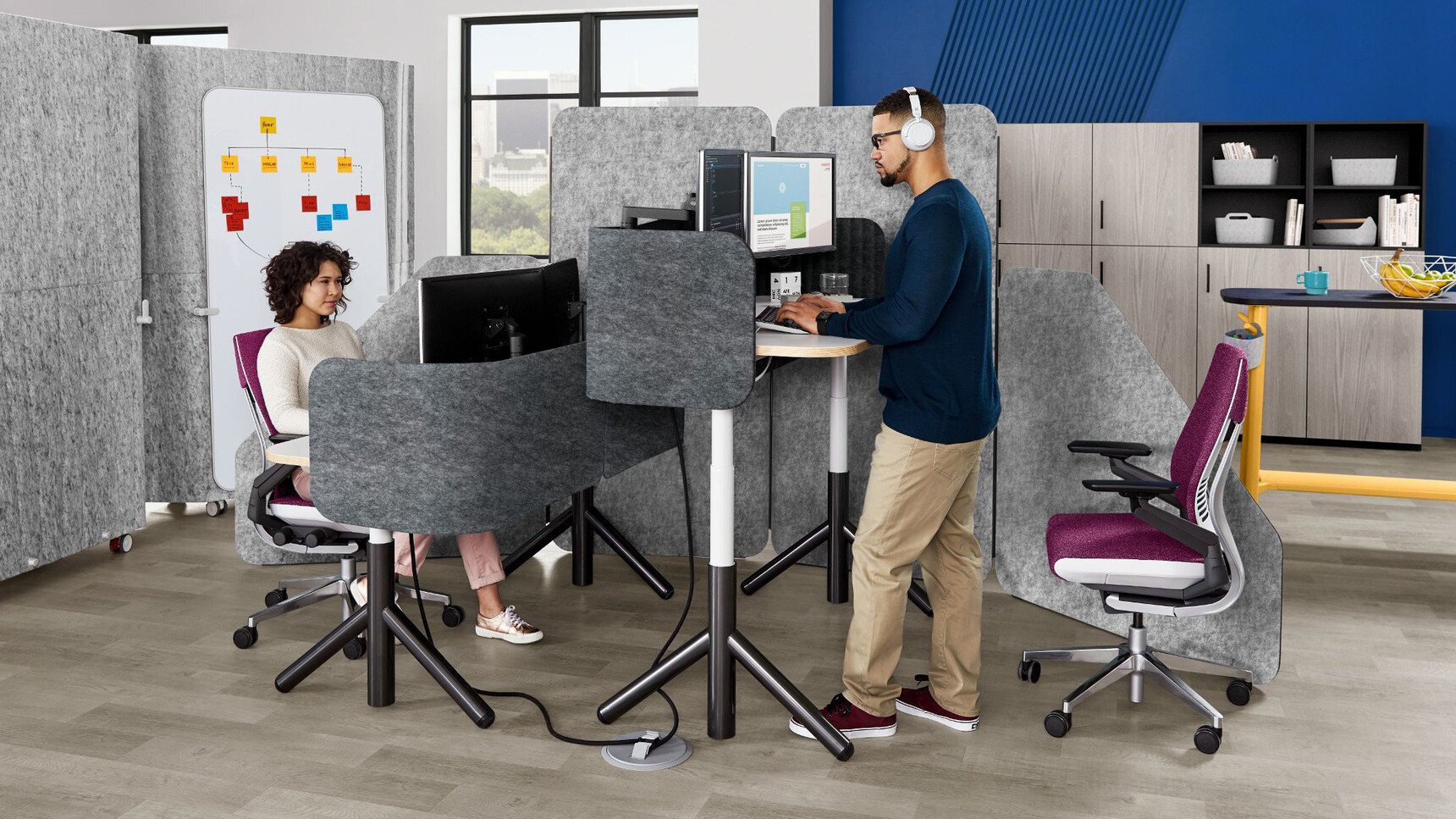 Two Steelcase Flex Height Adjustable Desks With White Table Top Finish And Black Leg Finish And Wood Edges With Dark Grey Privacy Screens With Two Gesture Wrapped Back Office Chairs In Use By Two People
