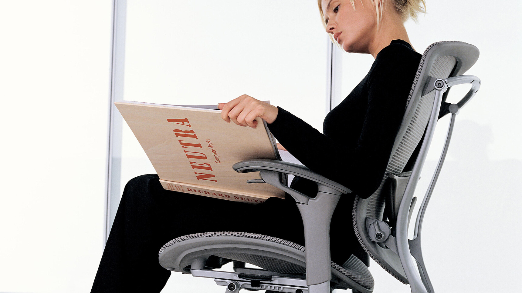 Okamura Contessa II Mesh Seat & Back Office Chair With Light Grey Cushion And Backrest With Grey Frame Finish With Grey 4-D Adjustable Arms In Use By A Person Reading A Book