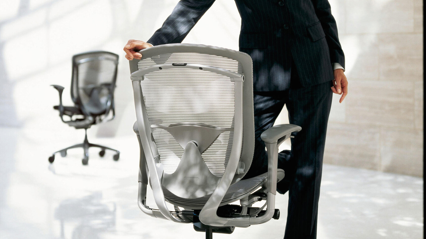 Two Okamura Contessa II Mesh Seat & Back Office Chairs With Light Grey Cushion And Backrest With Grey Frame Finish With Grey 4-D Adjustable Arms With A Person Leaning On The Chair