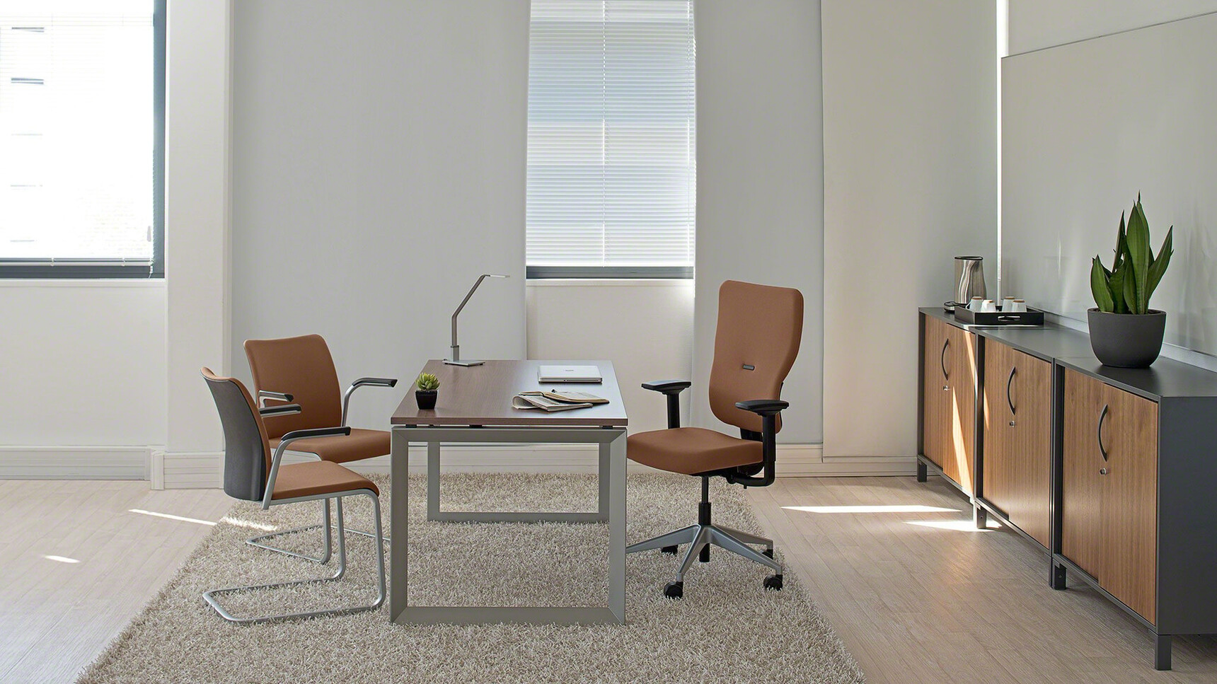Two Eastside Sled Base Meeting Chairs With Metal Base Finish Brown Upholstered Backrest Black Fixed Arms And Brown Upholstered Seat At Desk With Brown Steelcase Chair In Office Environment